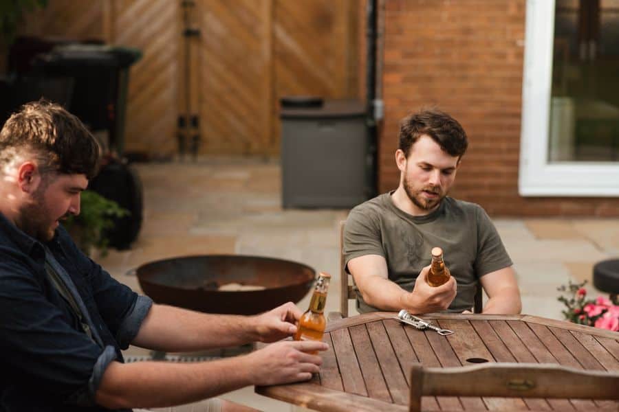 Two guys sitting on a table trying to remove the labels off a beer bottle