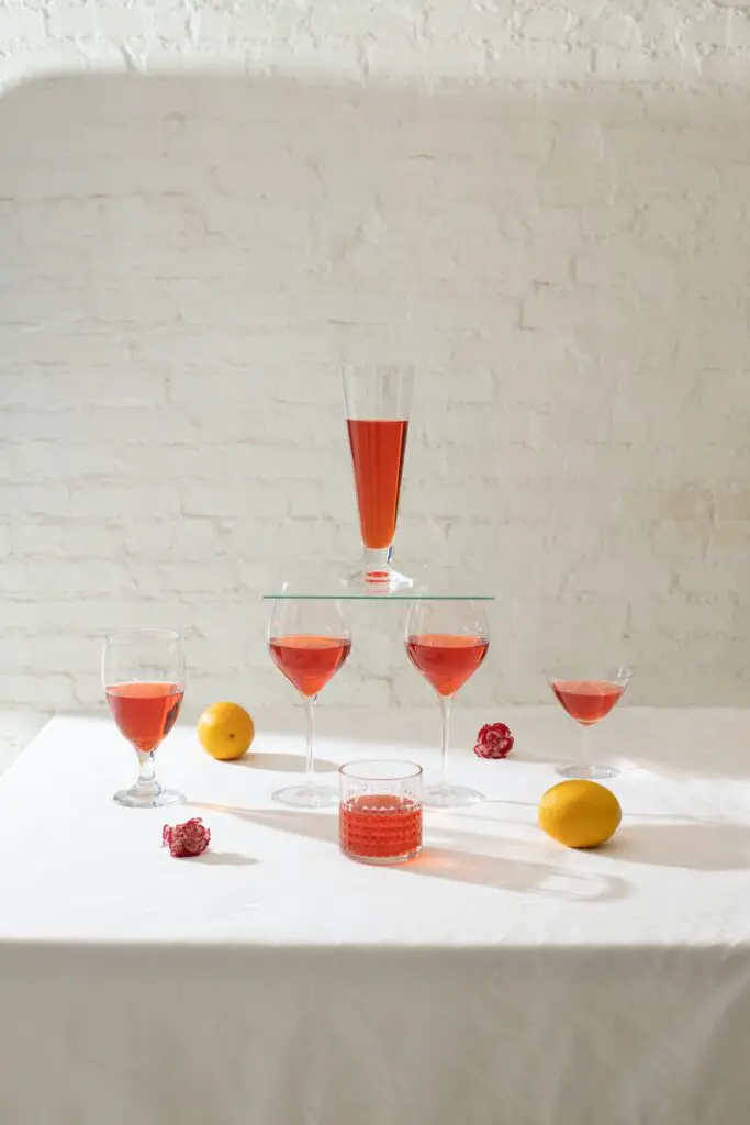 Different half-filled wine glasses near fresh oranges and pink flowers are arranged on top of a white tablecloth