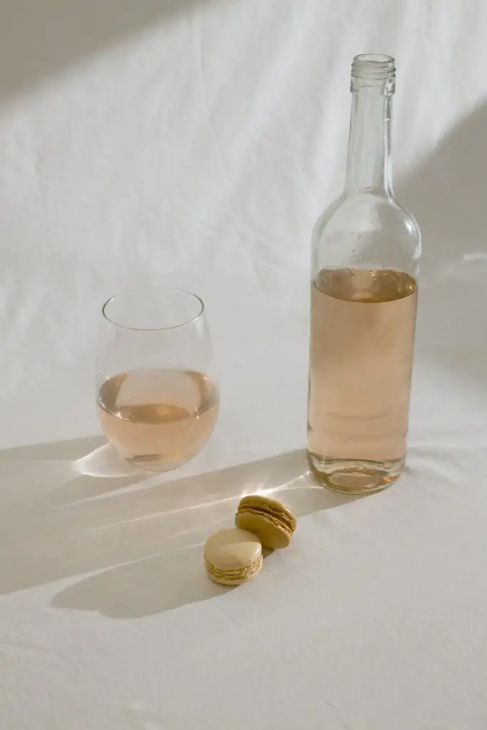 A wine bottle beside a clear glass half-filled with wine and macaroons is placed on a white surface