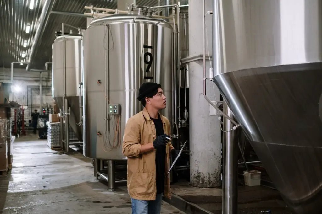 A man wearing a brown jacket, and black shirt is checking the metal still in a brewery