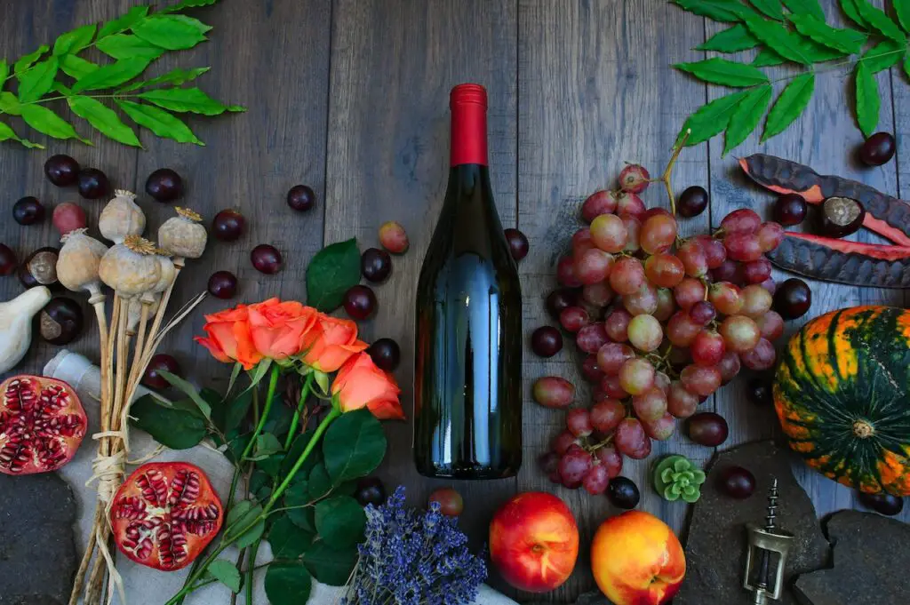 A black wine bottle surrounded by different fresh fruits and flowers on top of a brown wooden table