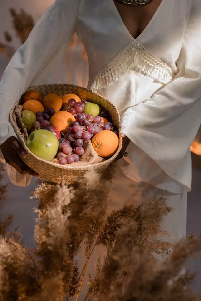 A woman holds a brown wicker basket filled with a variety of fruits while wearing a v-neck white long sleeves dress
