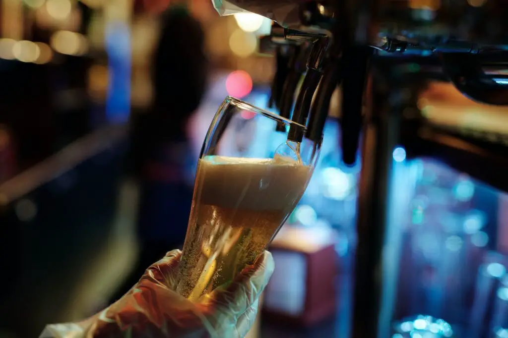 A person wearing plastic gloves holds a clear glass filling it with craft beer from a silver dispenser at the bar