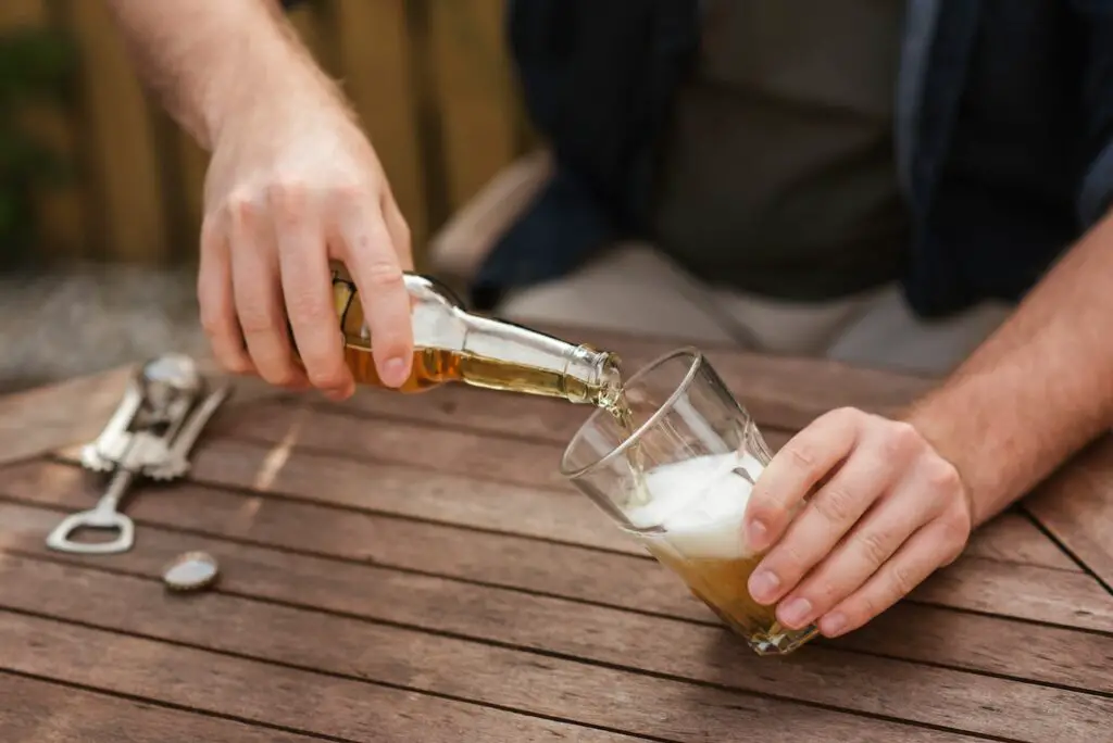 A man pouring a foamy beer in clear glass and a bottle opener is placed on a brown wooden table