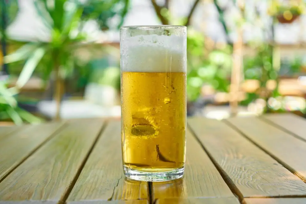 A clear glass filled with cold foamy beer was placed on a brown wooden table outside