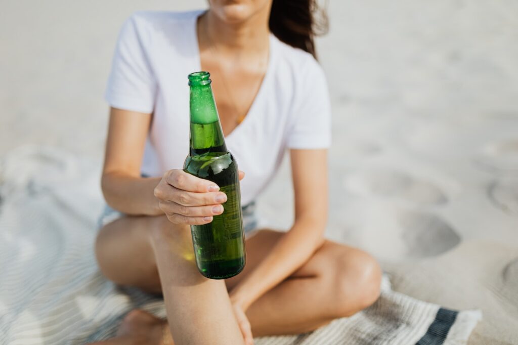 A woman in a white t-shirt holds a green bottle of beer while seated on a blanket placed on the sand
