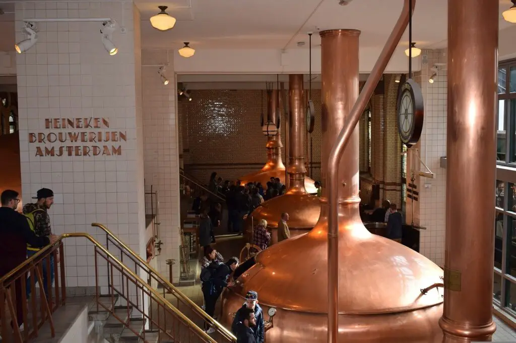 People checking the copper tanks that are lined up near a white tiled wall near golden railings in a brewery