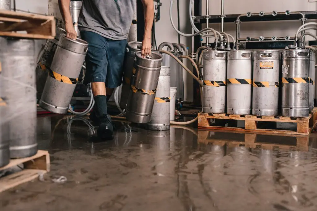 A person in a t-shirt and blue shorts holding stainless steel kegs at a brewery