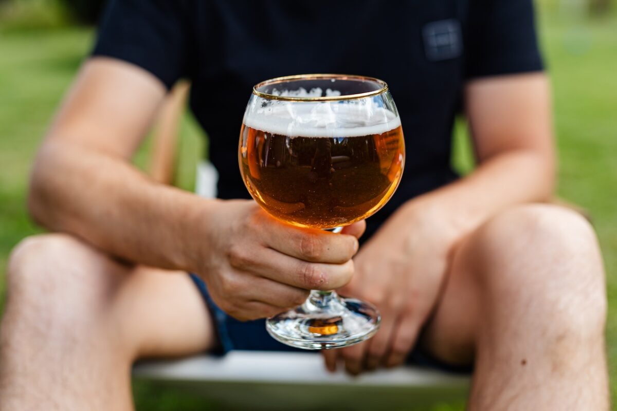 A man in a black t-shirt and shorts holds a clear glass filled with foamy beer