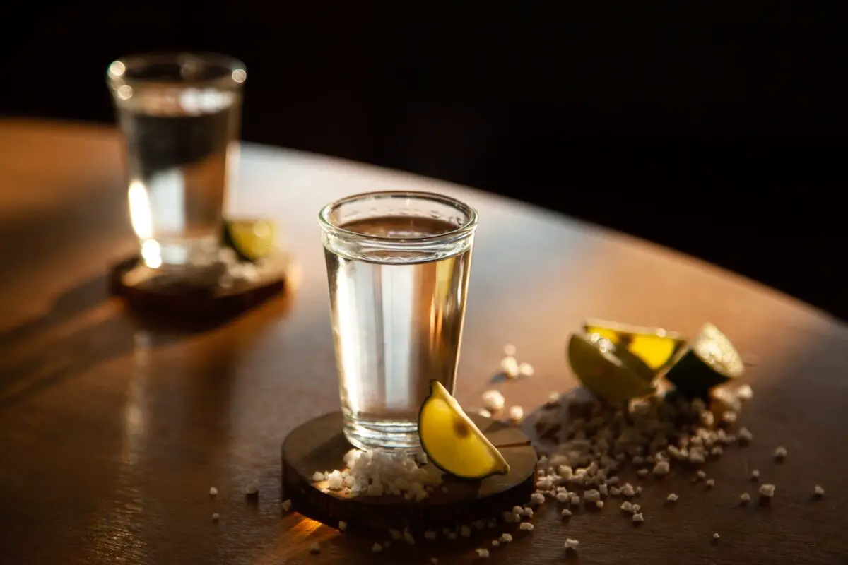 Two shots of tequila beside salt and lemon placed on a wooden coaster